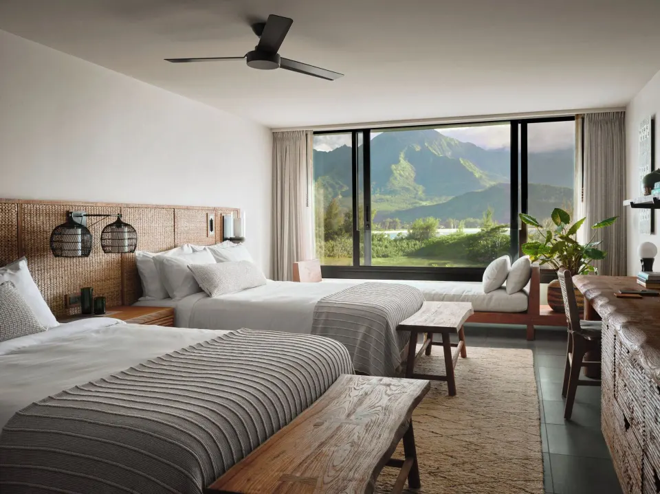 Two queen beds and a window bench overlook green mountains at 1 Hotel Hanalei Bay.