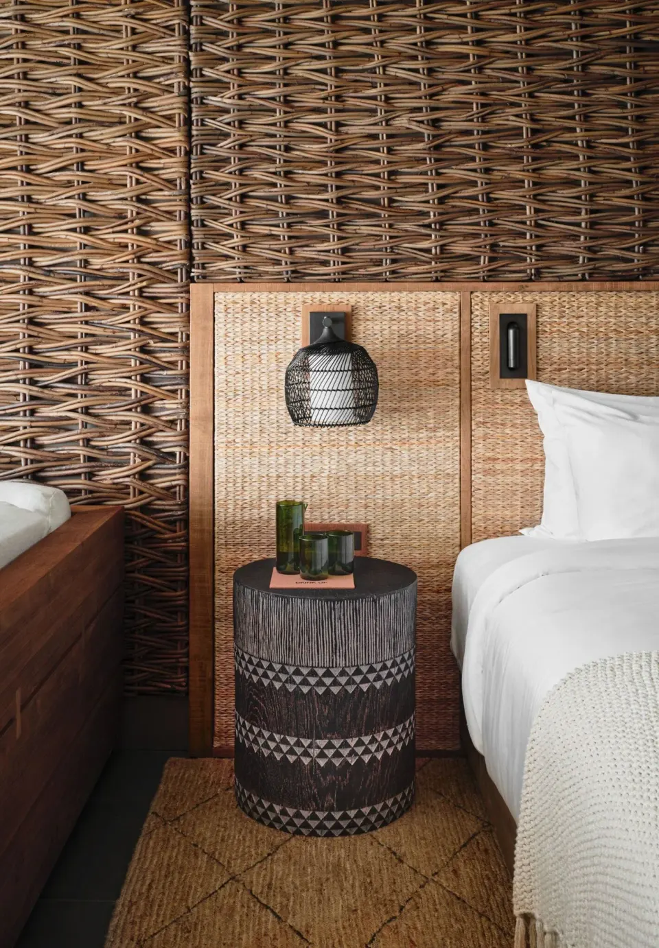 Guest room bed at 1 Hotel Hanalei Bay with woven wall panels, bedside table and white linens.