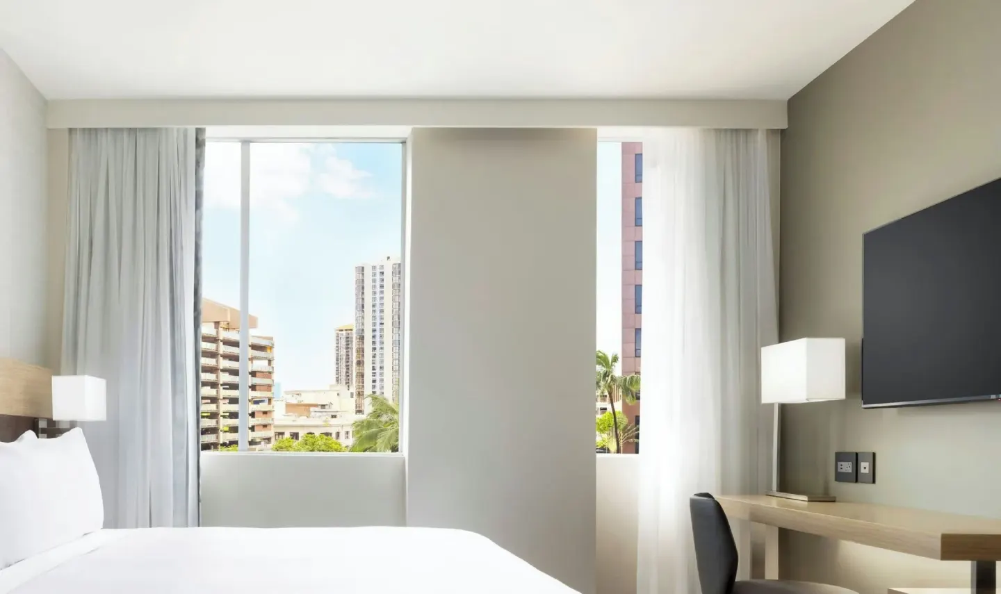 Guest room bed, desk and wall-mounted TV facing city windows at AC Hotel Honolulu.