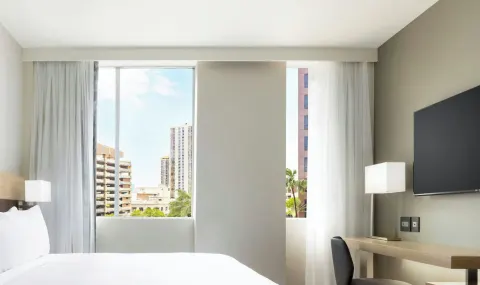 Guest room bed, desk and wall-mounted TV facing city windows at AC Hotel Honolulu.