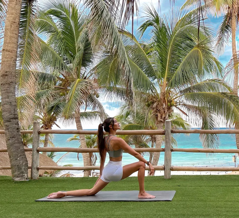 Woman doing a lunge on a yoga mat with palm trees and turquoise sea at Ahau Tulum.
