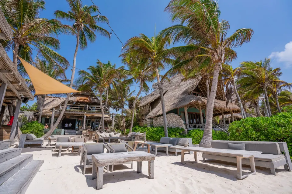 Sunlit beach loungers and tables beneath palm trees at Ahau Tulum.
