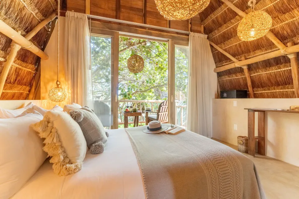 Deluxe Room at Ahau Tulum with palapa roof, king bed and balcony visible through glass doors.