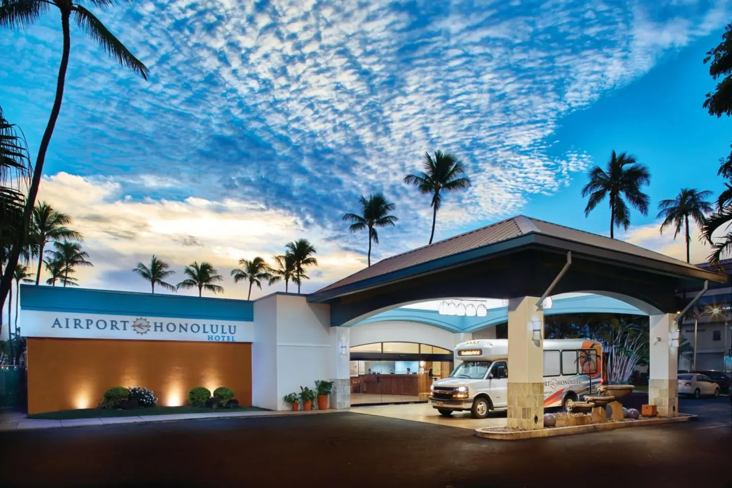 Evening exterior of Airport Honolulu Hotel with lit entrance canopy and shuttle van.
