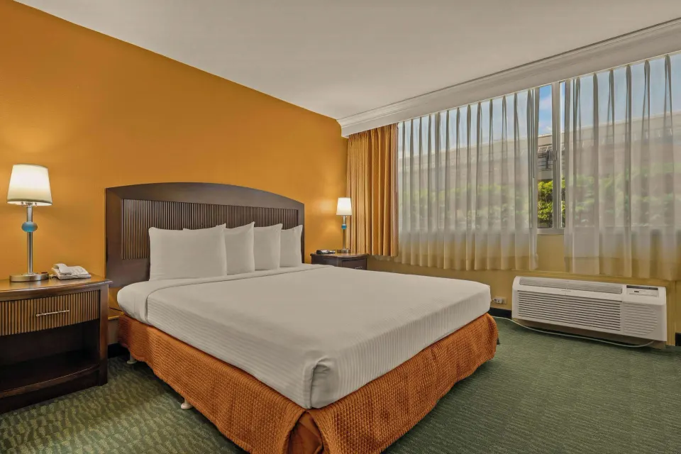 King bed beside a large curtained window and wall air unit at Airport Honolulu Hotel.
