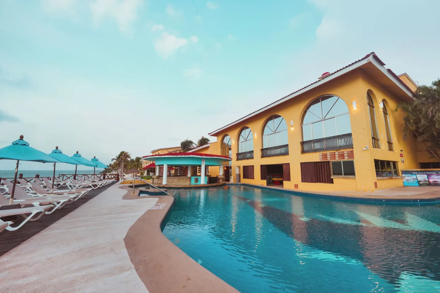 Seaside pool with blue umbrellas and arched-window building at All Ritmo Cancun Resort & Water Park.
