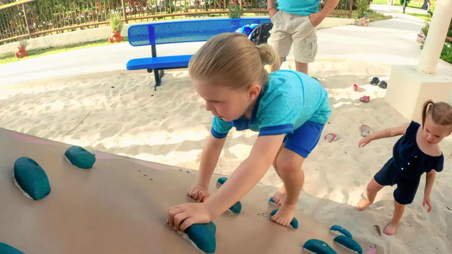 Children climb on a shaded play wall in a sandy kids area at Allegro Cozumel.