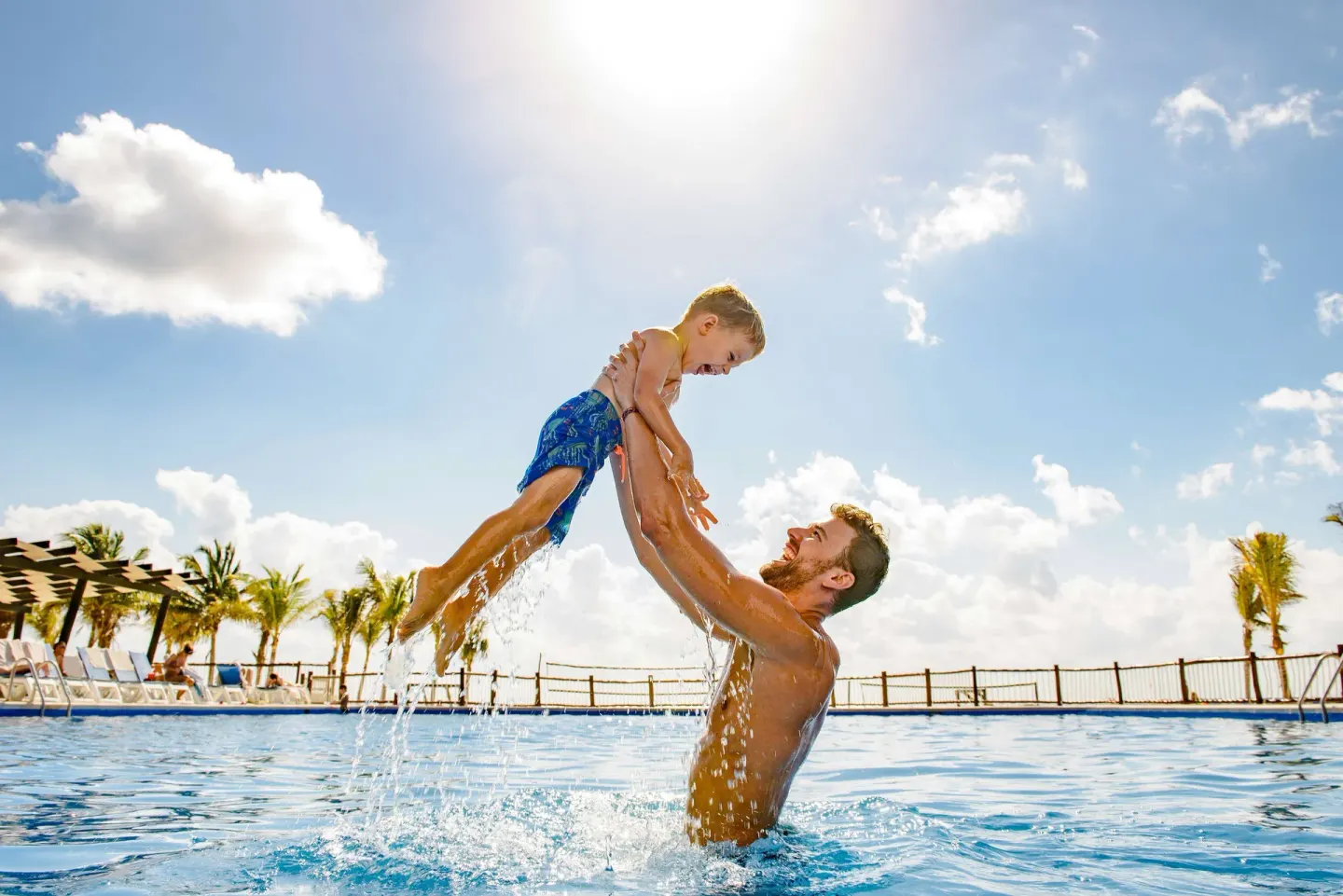 A man lifts a child from the pool water at Allegro Playacar under a bright sky.