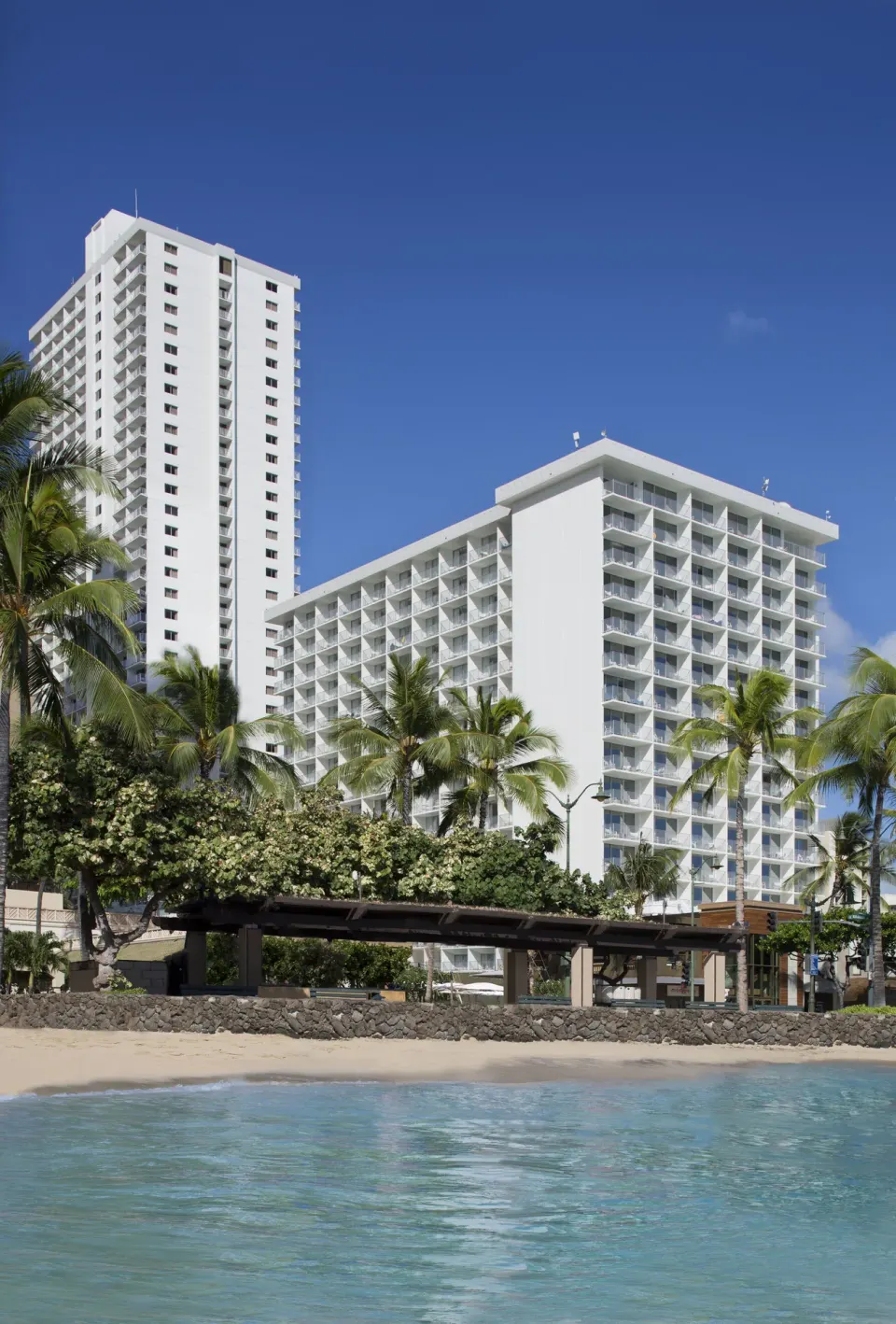 Beachfront hotel towers rise behind palms and water at 'Alohilani Resort Waikiki Beach.