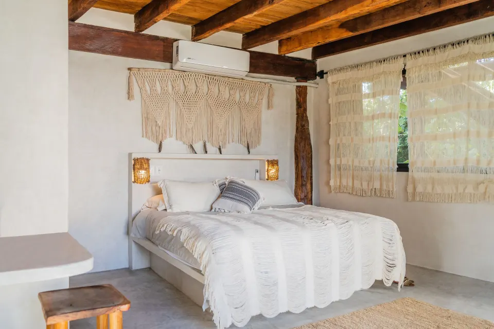Bright bed beneath macramé wall hanging and wood beams at Amansala