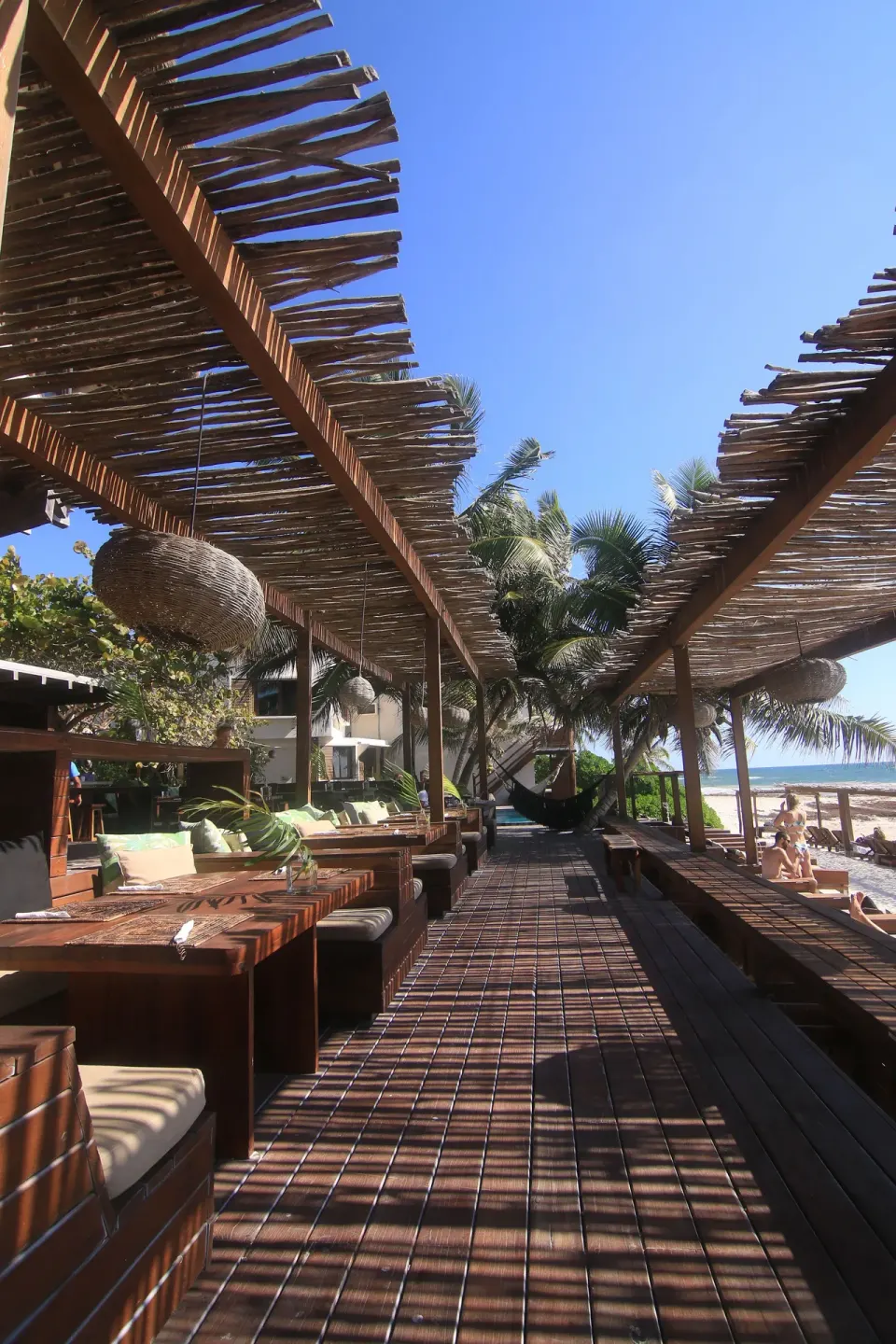 Shaded wooden walkway with cushioned booths and hanging lanterns by the beach at Amansala