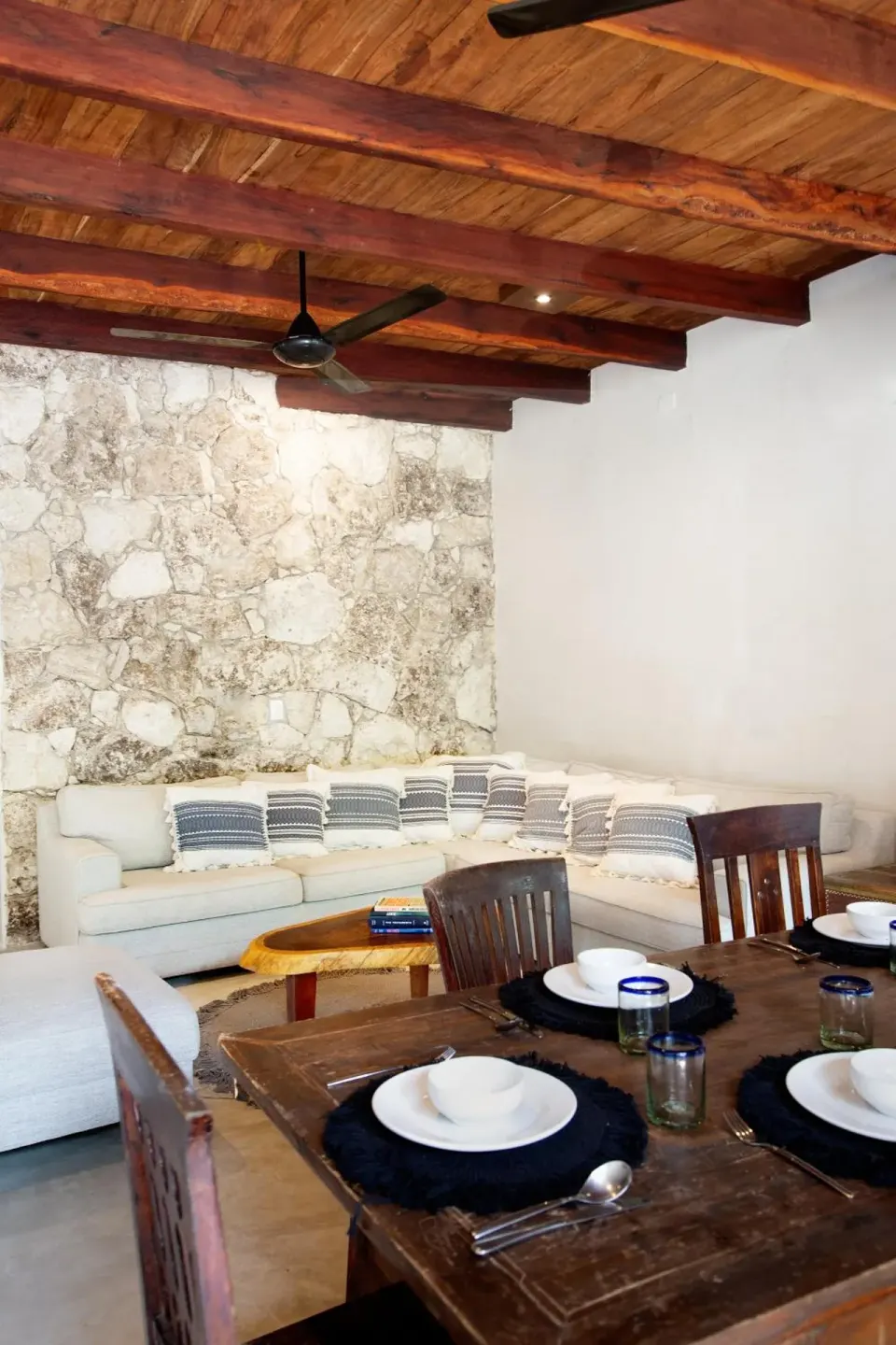 Dining table set for four beside a white sectional sofa and textured stone wall at Amansala