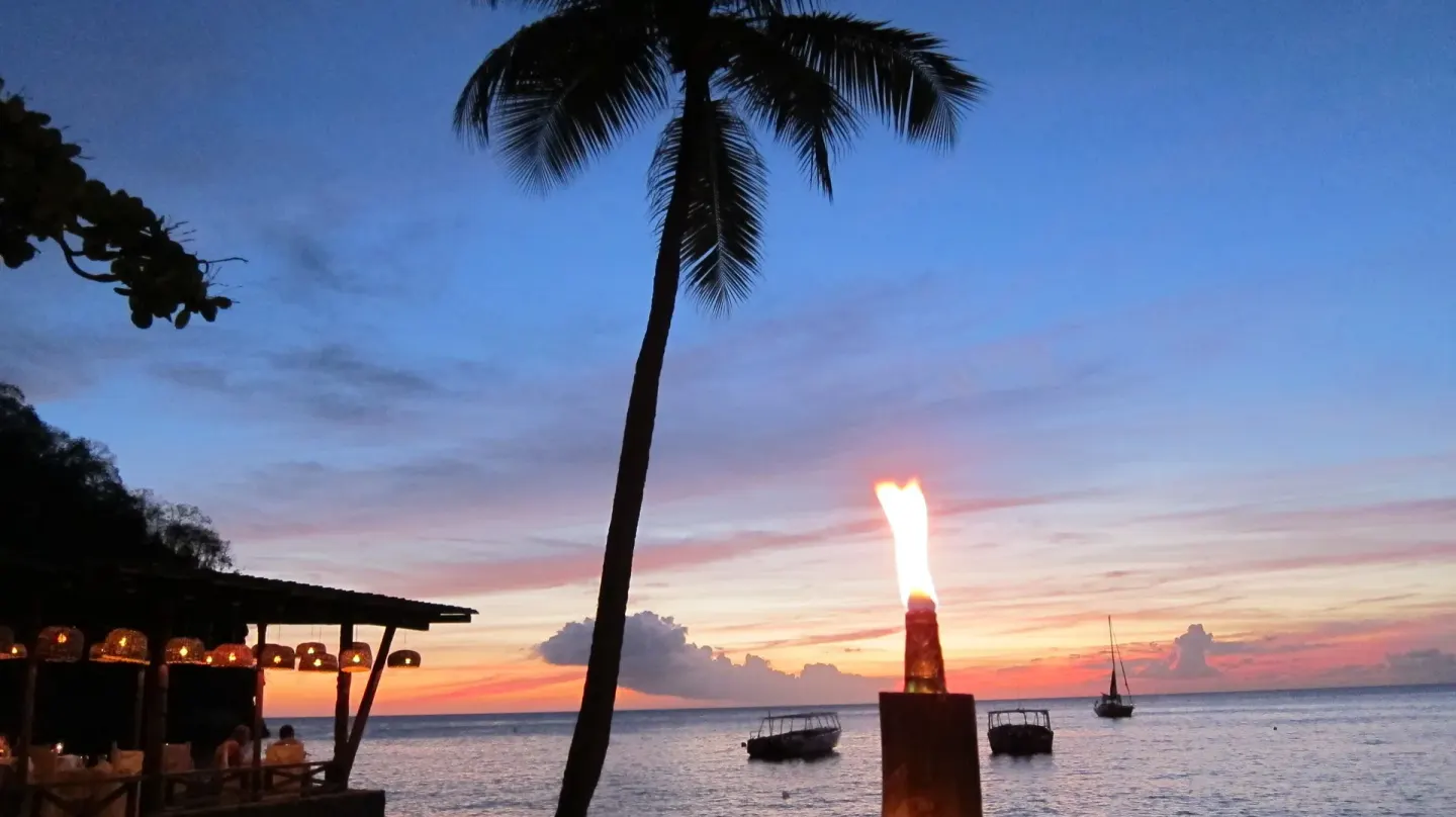 Sunset beach scene at Anse Chastanet Resort with a torch, palm tree, and boats offshore.