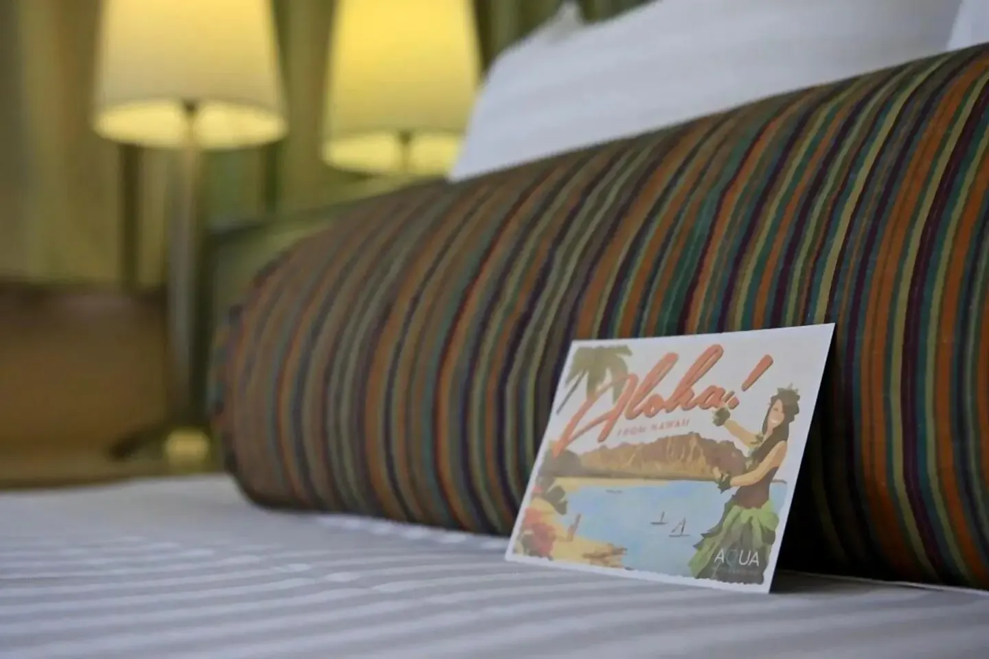 Junior Suite bed detail with striped bolster pillow and bedside lamps at Aqua Aloha Surf Waikiki.