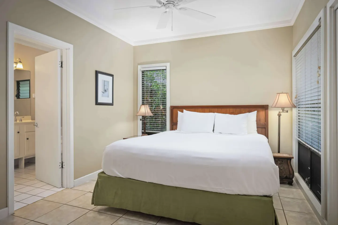 Guest bedroom with king bed, bedside lamps, blinds, and open bath door at Aston at Poipu Kai.