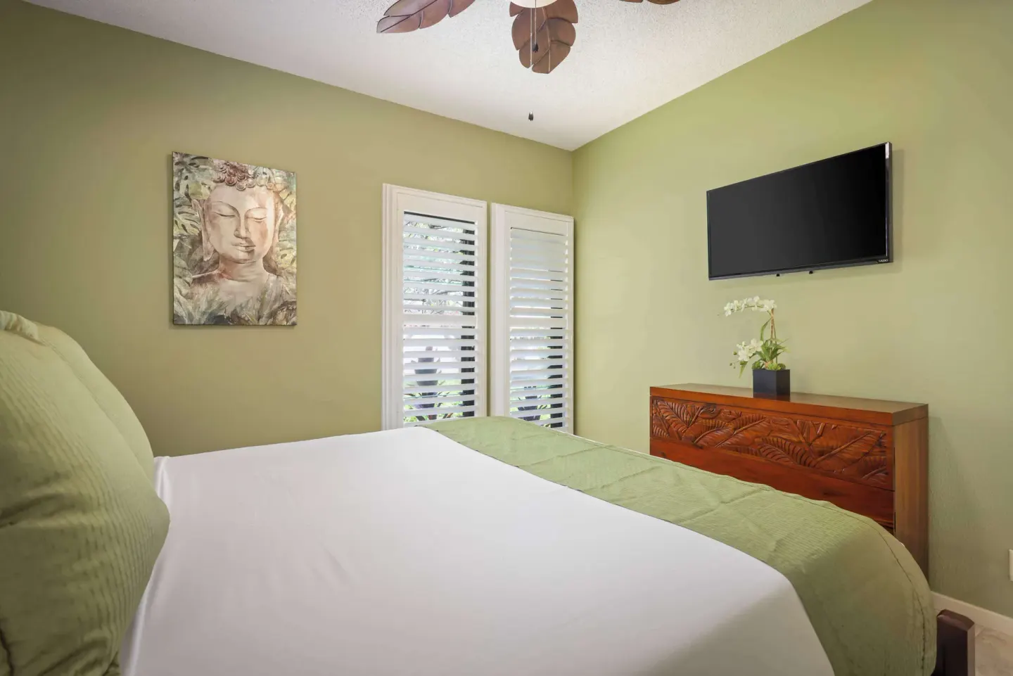 Guest bedroom bed facing a dresser, shutters, and wall-mounted TV at Aston at Poipu Kai.
