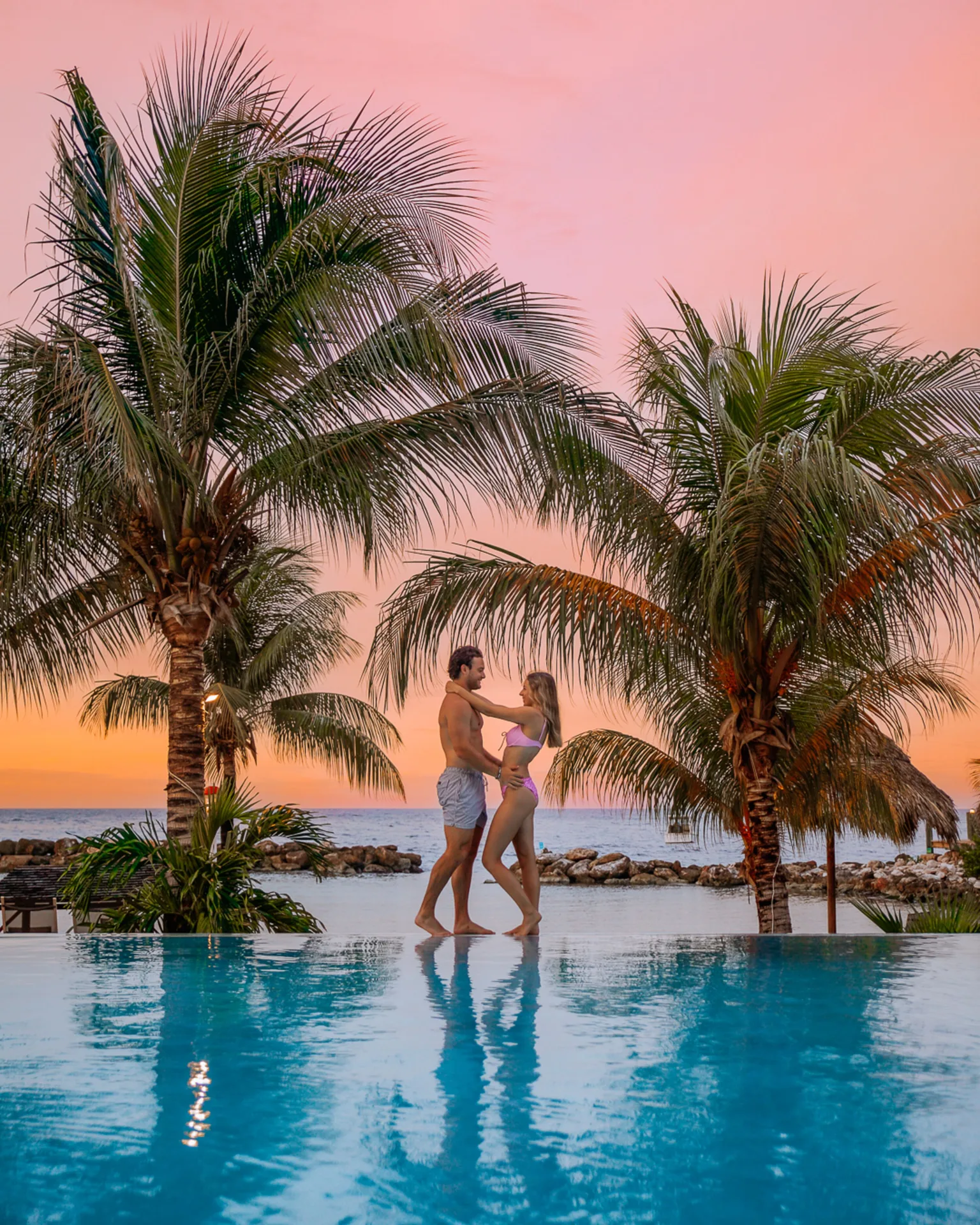 Couple standing by the pool between palms at sunset at Avila Beach Hotel.