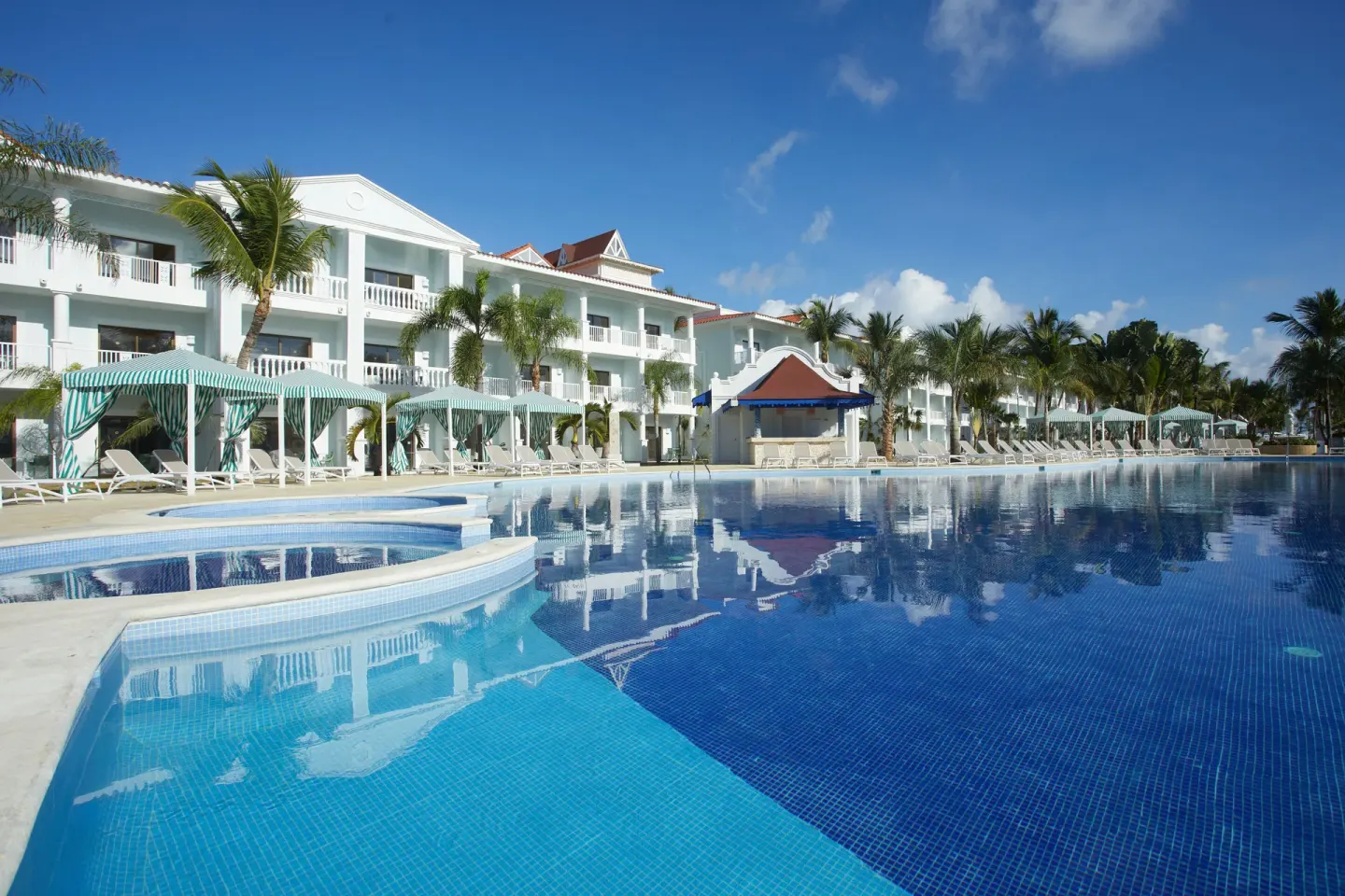 Blue pool edge with cabanas and white buildings at Bahia Principe Explore Esmeralda.