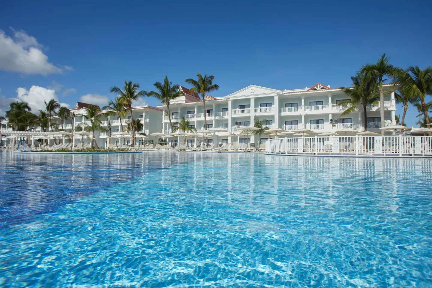 Low pool-level view of blue water, palms and guest buildings at Bahia Principe Explore Esmeralda.