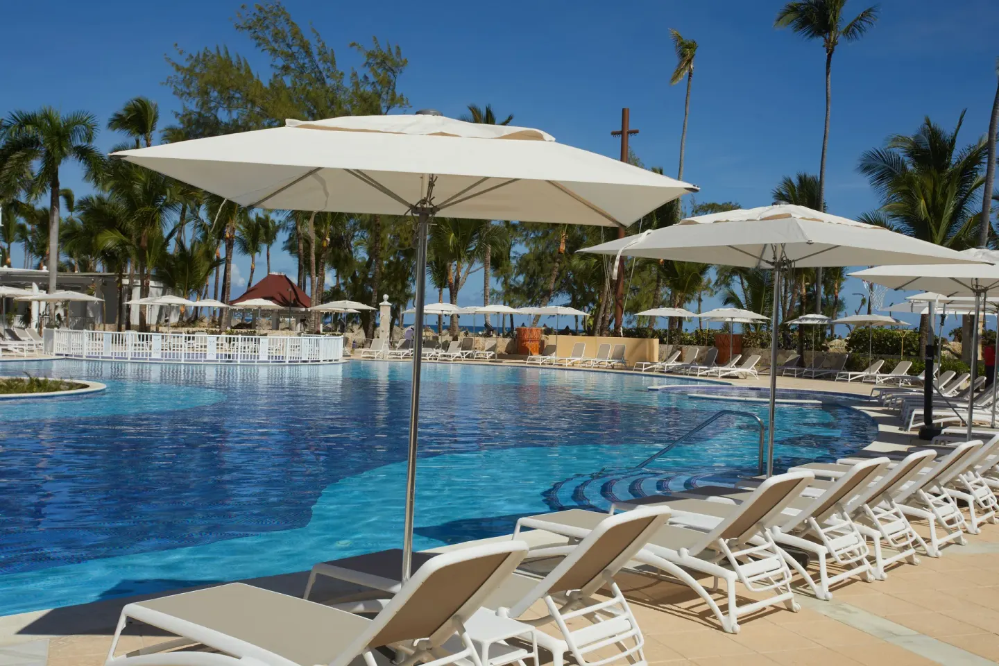 Pool loungers under large white umbrellas beside palm trees at Bahia Principe Explore Esmeralda.