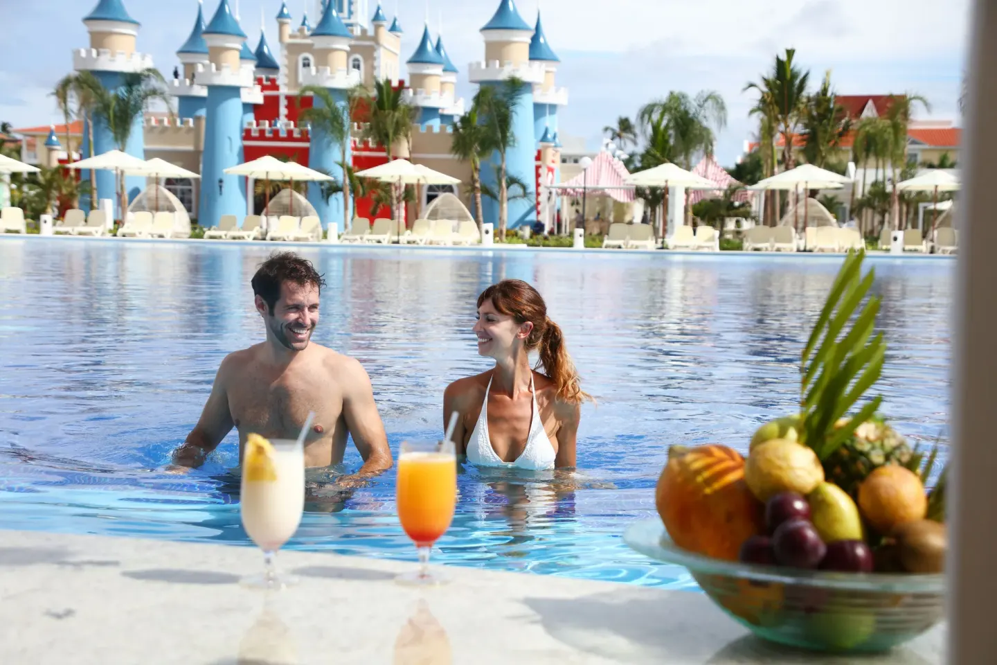 Two guests in the pool with drinks and a castle backdrop at Bahia Principe Explore Legend.
