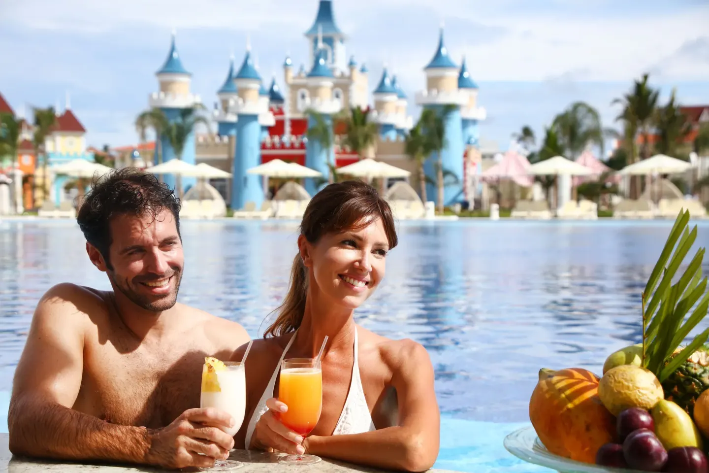 Couple holding poolside drinks with castle towers behind at Bahia Principe Explore Legend.