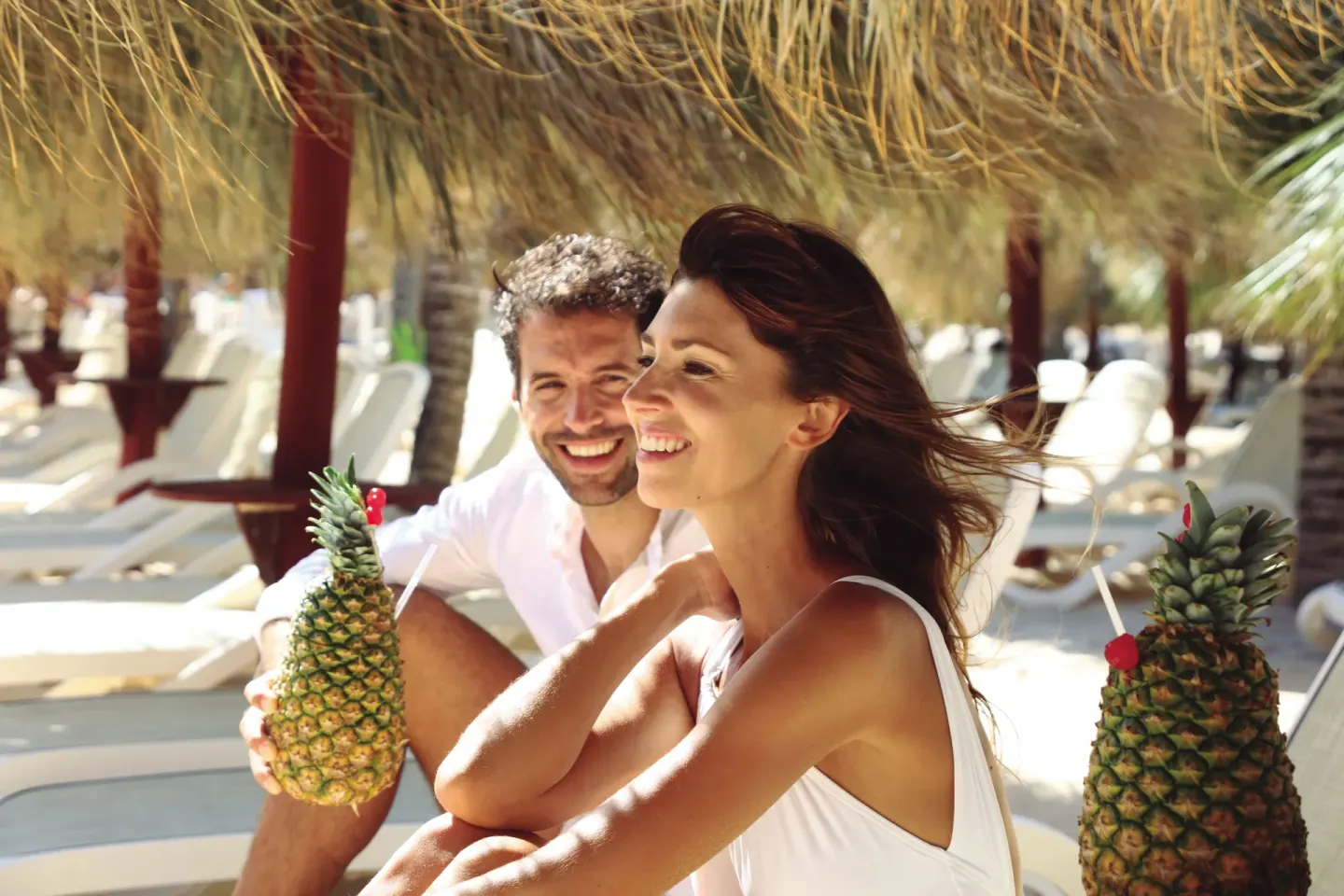 Couple holding pineapple drinks under a beach palapa at Bahia Principe Grand Aquamarine.