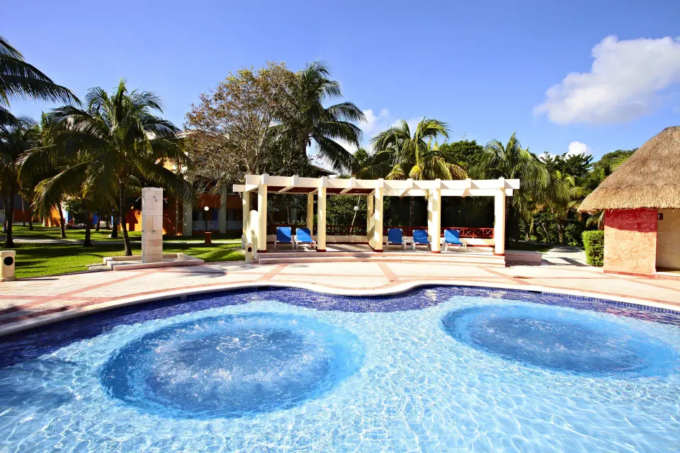 Large freeform resort pool surrounded by palm trees and thatched roofs at Bahia Principe Grand Coba