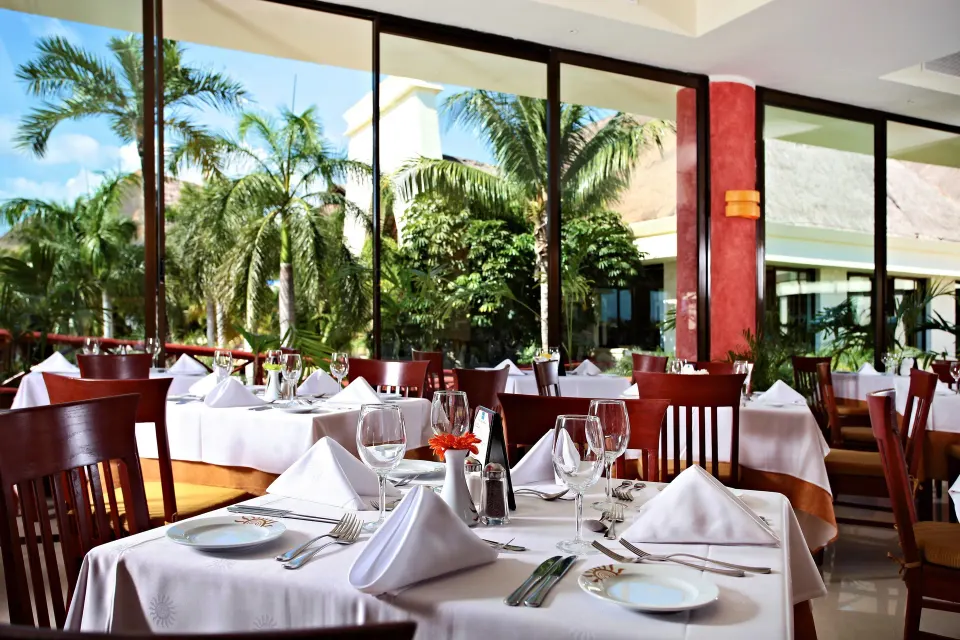 Sunlit restaurant dining tables near floor-to-ceiling windows at Bahia Principe Grand Coba