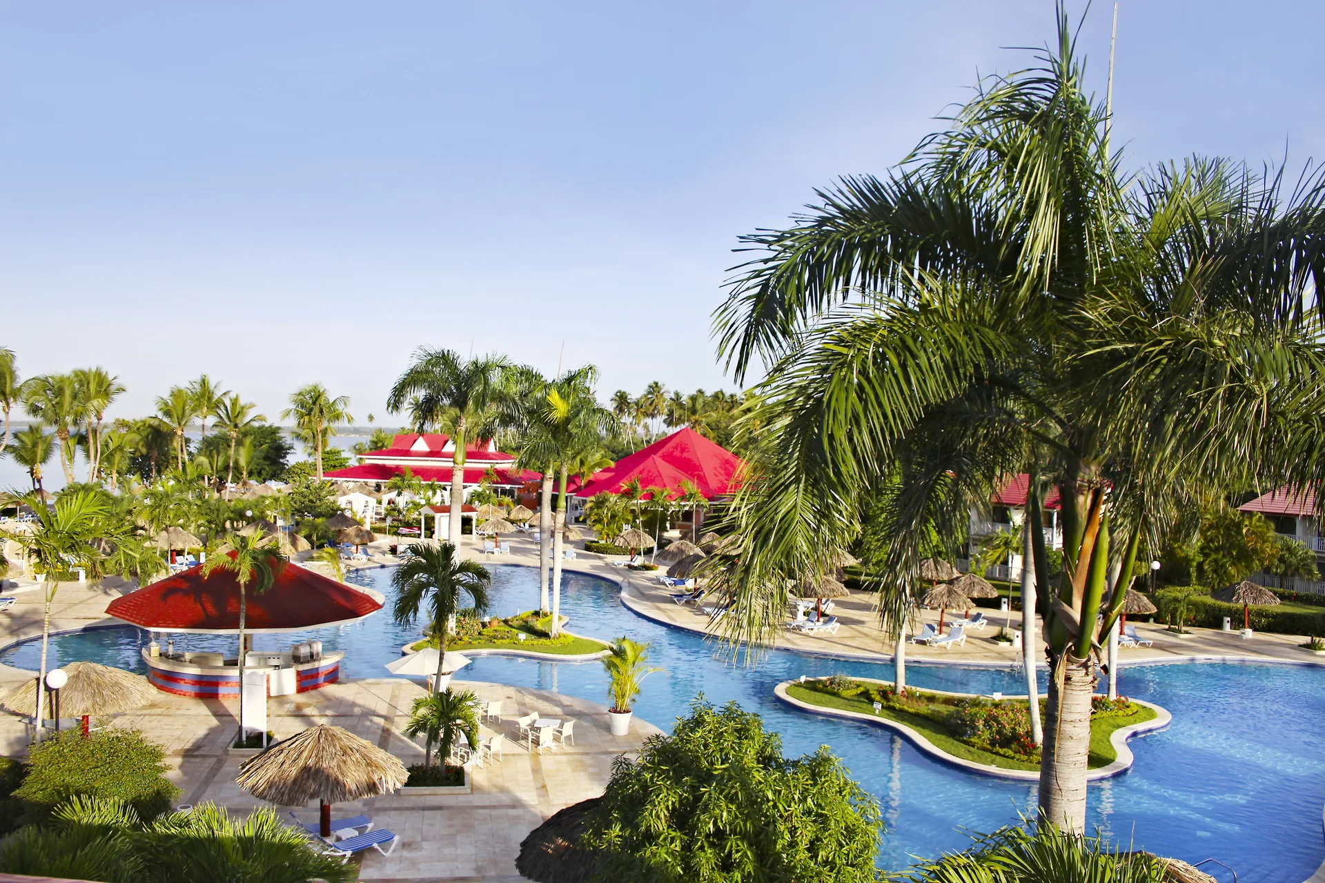 Curving pool at Bahia Principe Grand La Romana with palm islands, palapas, loungers and red-roof structures.