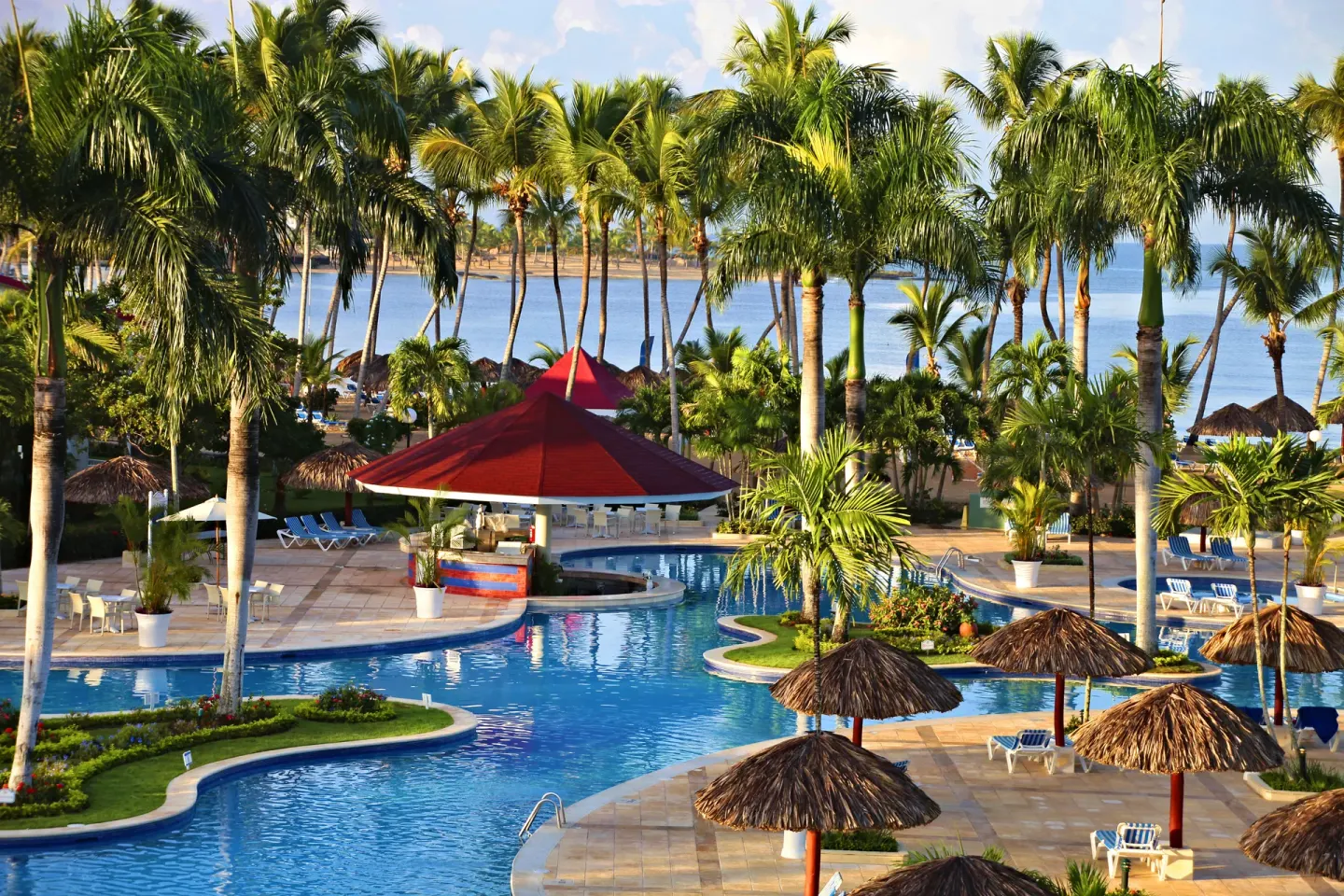 Pool area at Bahia Principe Grand La Romana with palms, palapas, loungers and a red-roof swim-up bar.
