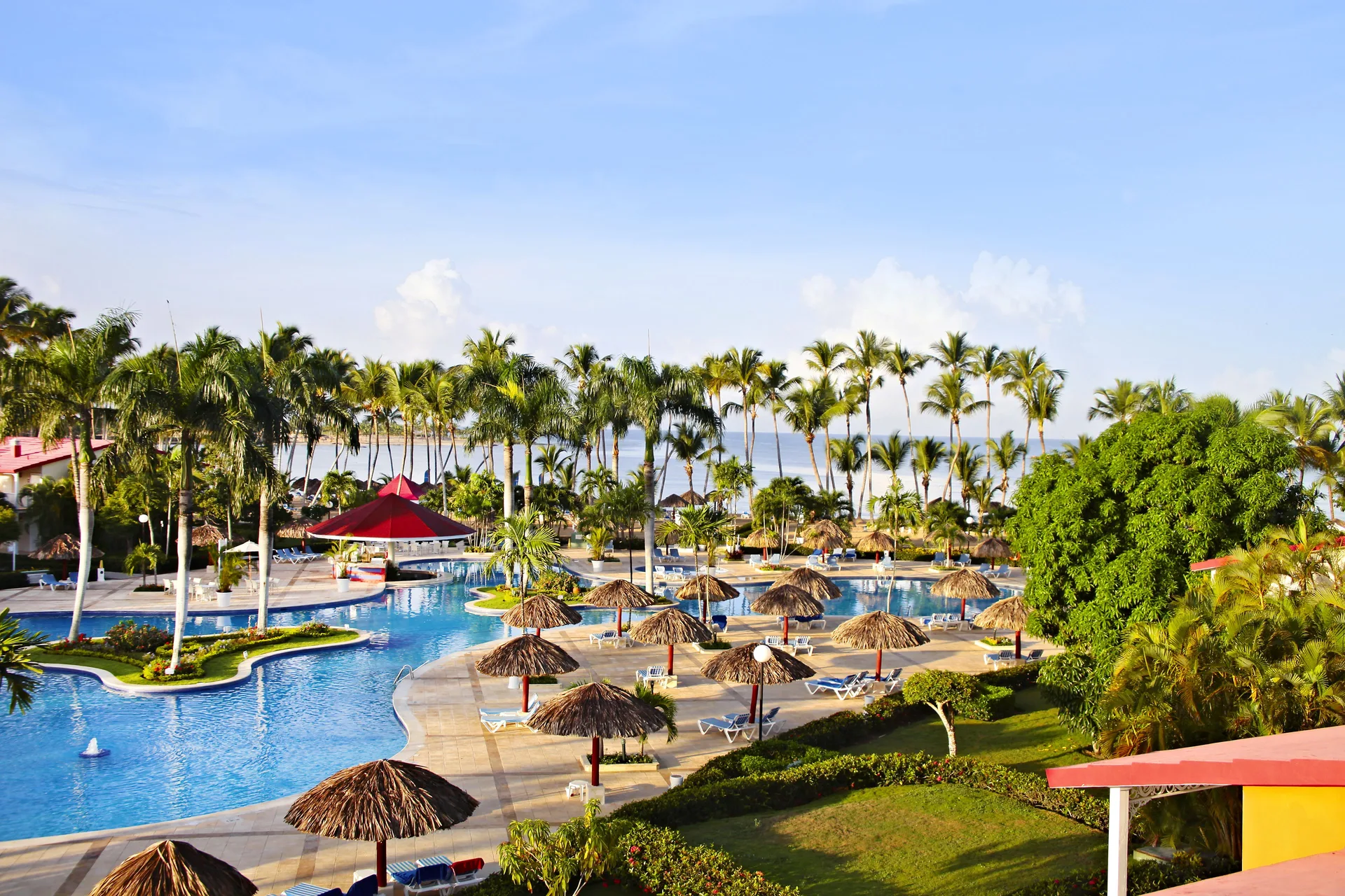 Wide pool view at Bahia Principe Grand La Romana with palapas, loungers, palms and the sea beyond.