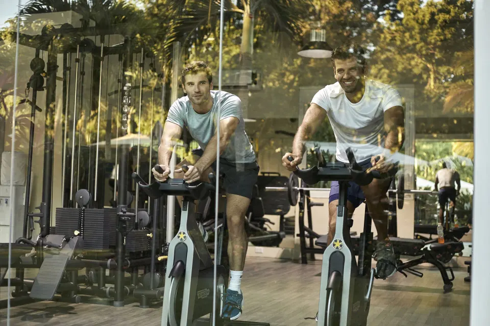 Two men cycling on stationary bikes in a fitness room at Bahia Principe Grand Tulum