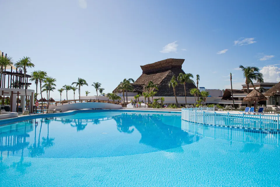 Blue resort pool with palm trees and thatched-roof buildings at Bahia Principe Grand Tulum