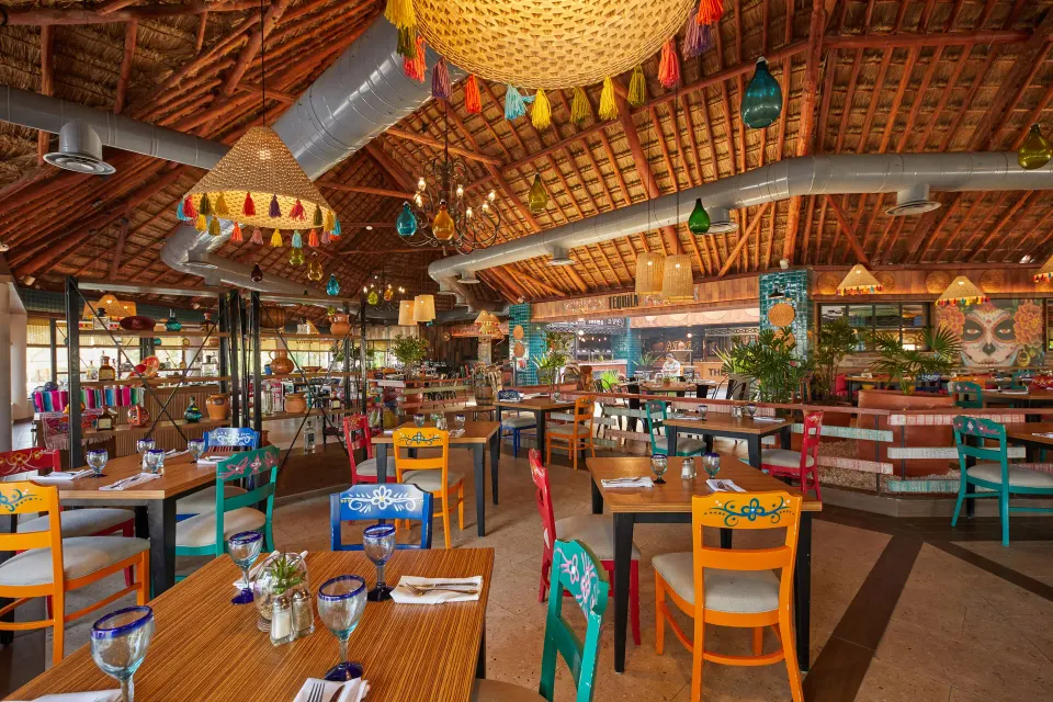 Colorful open dining room with bright painted chairs and large woven chandeliers at Bahia Principe Grand Tulum