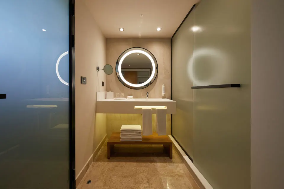 Long view down a bathroom with frosted-glass partitions and lit round mirror at Bahia Principe Luxury Akumal