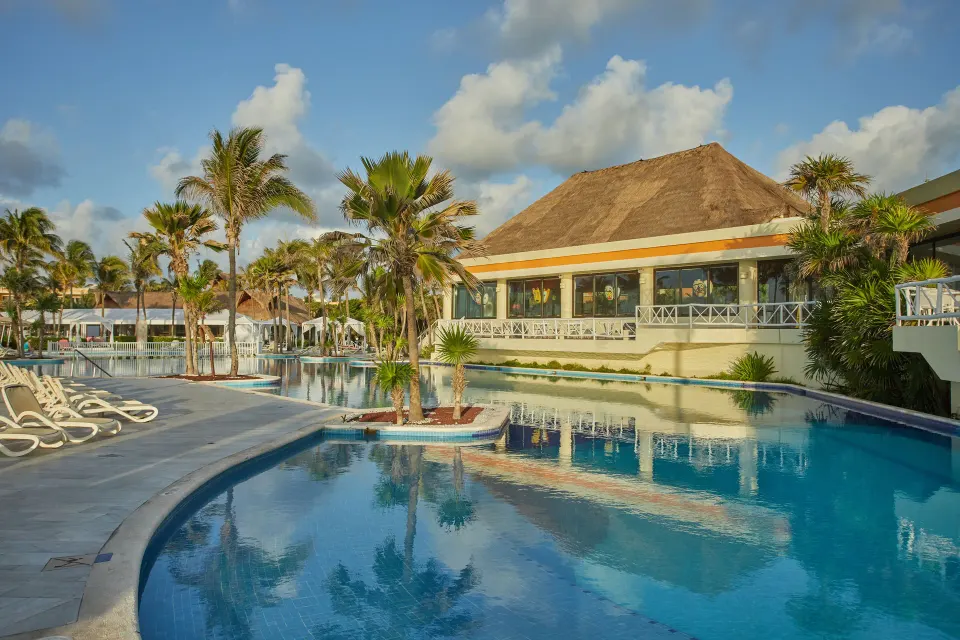 Curved outdoor resort pool with palm trees and a thatched-roof building at Bahia Principe Luxury Akumal