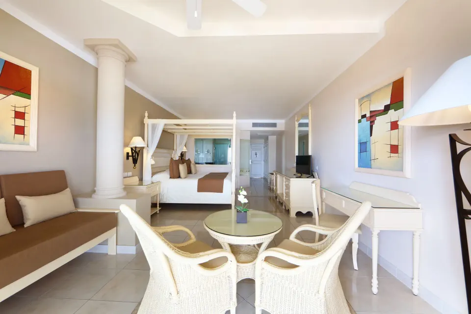 Long view of a junior suite with canopy bed, wicker dining set and neutral decor at Bahia Principe Luxury Akumal