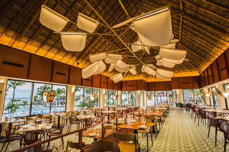 Long open dining hall with thatched roof and sculptural light fixtures at Bahia Principe Luxury Akumal