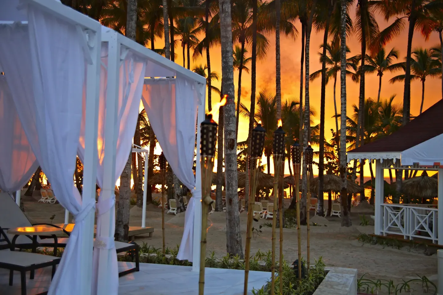 Beach cabanas, palm trees and tiki torches at sunset at Bahia Principe Luxury Bouganville.
