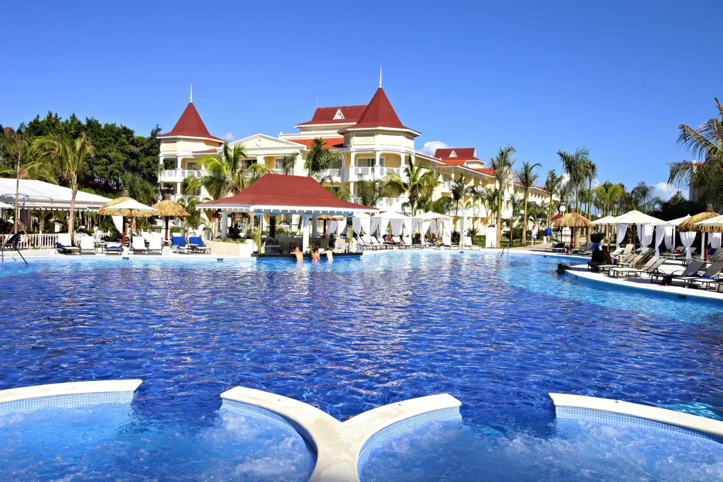 Large blue pool with loungers and red-roofed resort buildings at Bahia Principe Luxury Bouganville.