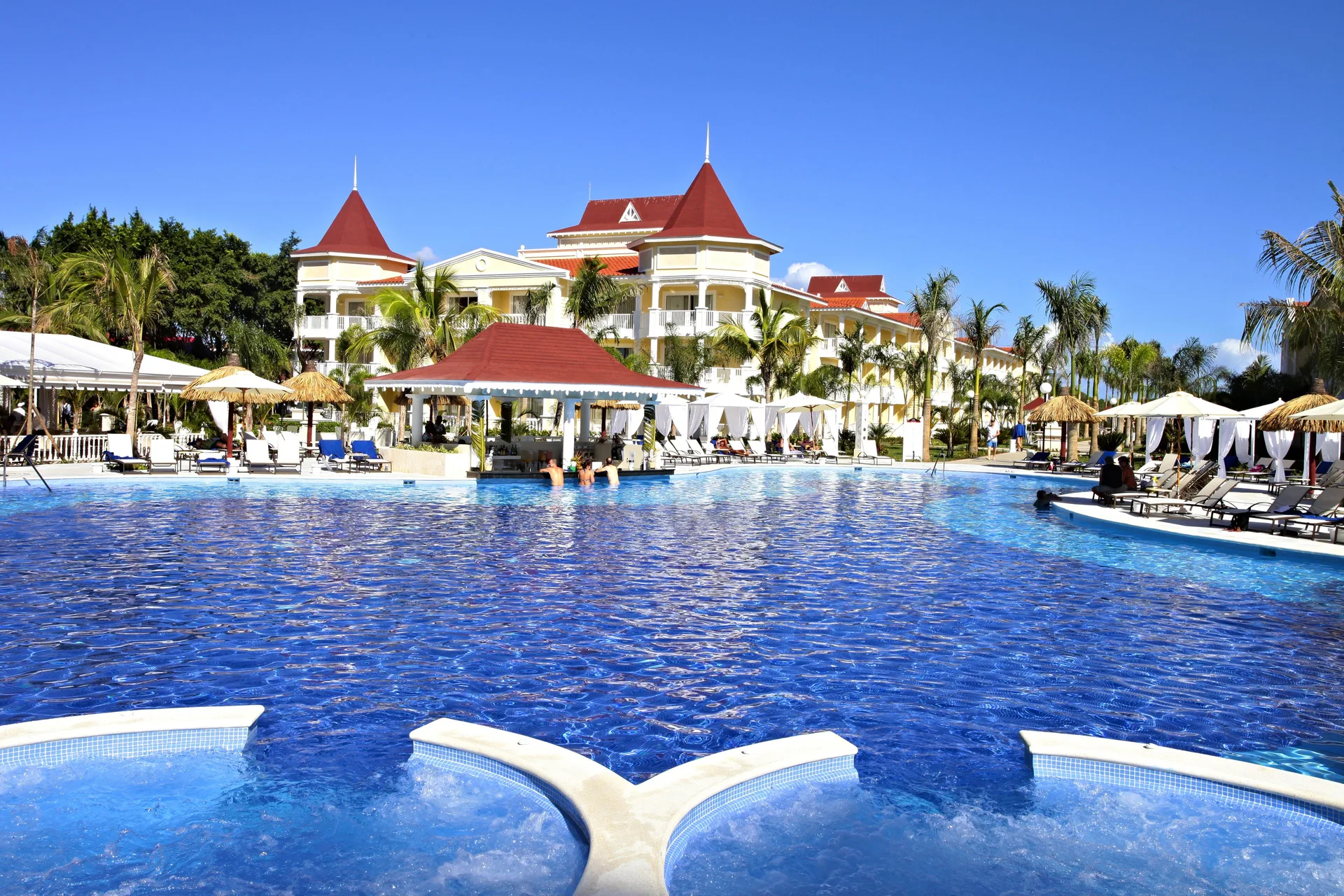 Large blue pool with loungers and red-roofed resort buildings at Bahia Principe Luxury Bouganville.