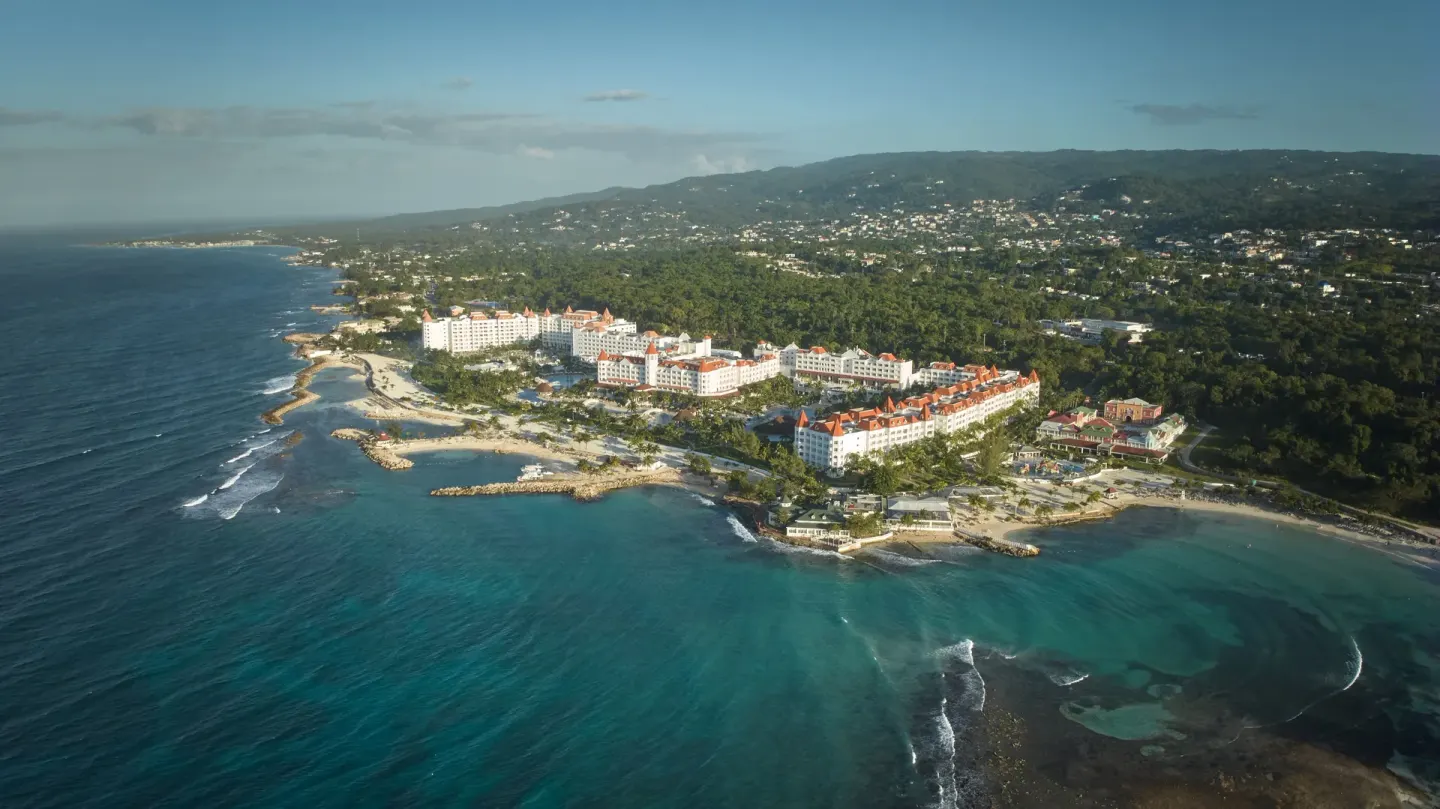 Wide aerial view of Bahia Principe Luxury Runaway Bay on the coast below green hills.