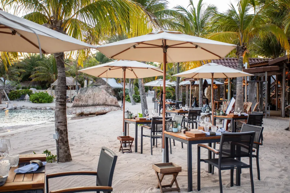 Beachfront dining tables with umbrellas on sand beside the lagoon at Baoase Luxury Resort