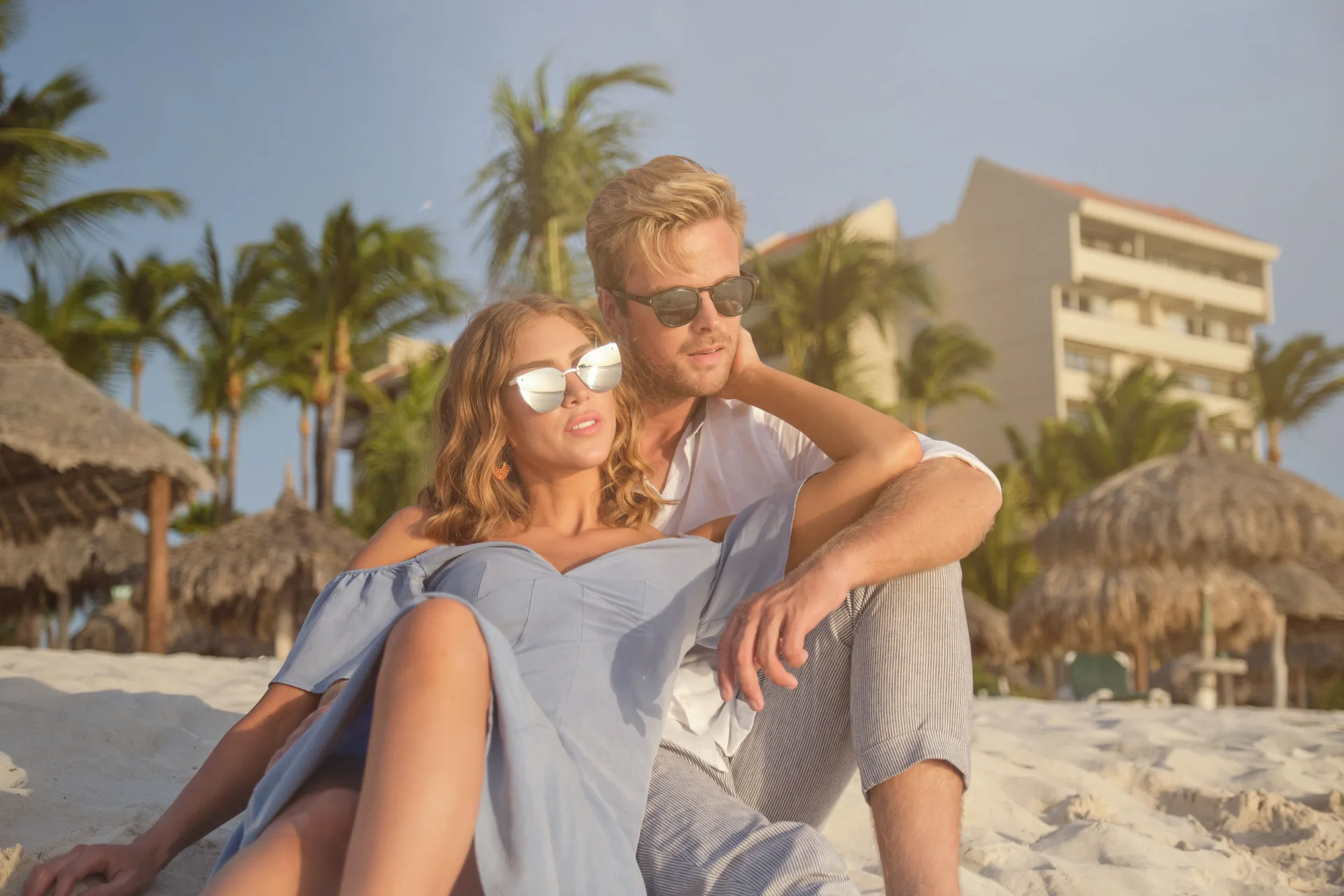 Couple seated on the sandy beach with palapas and resort buildings behind them at Barceló Aruba.