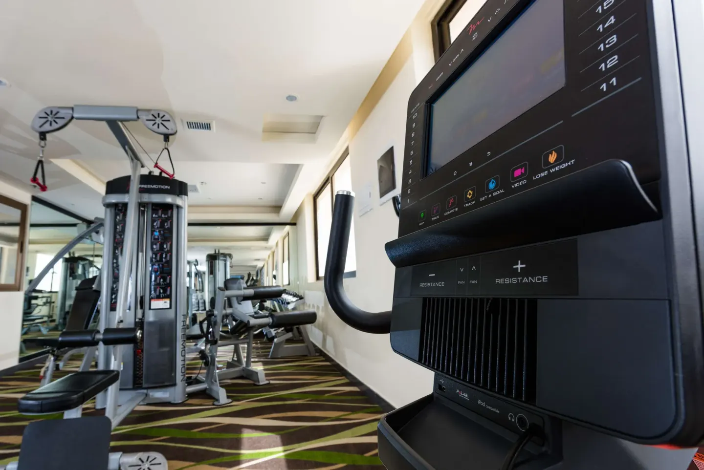 Close-up of an exercise machine console with gym equipment behind it at Barceló Aruba.