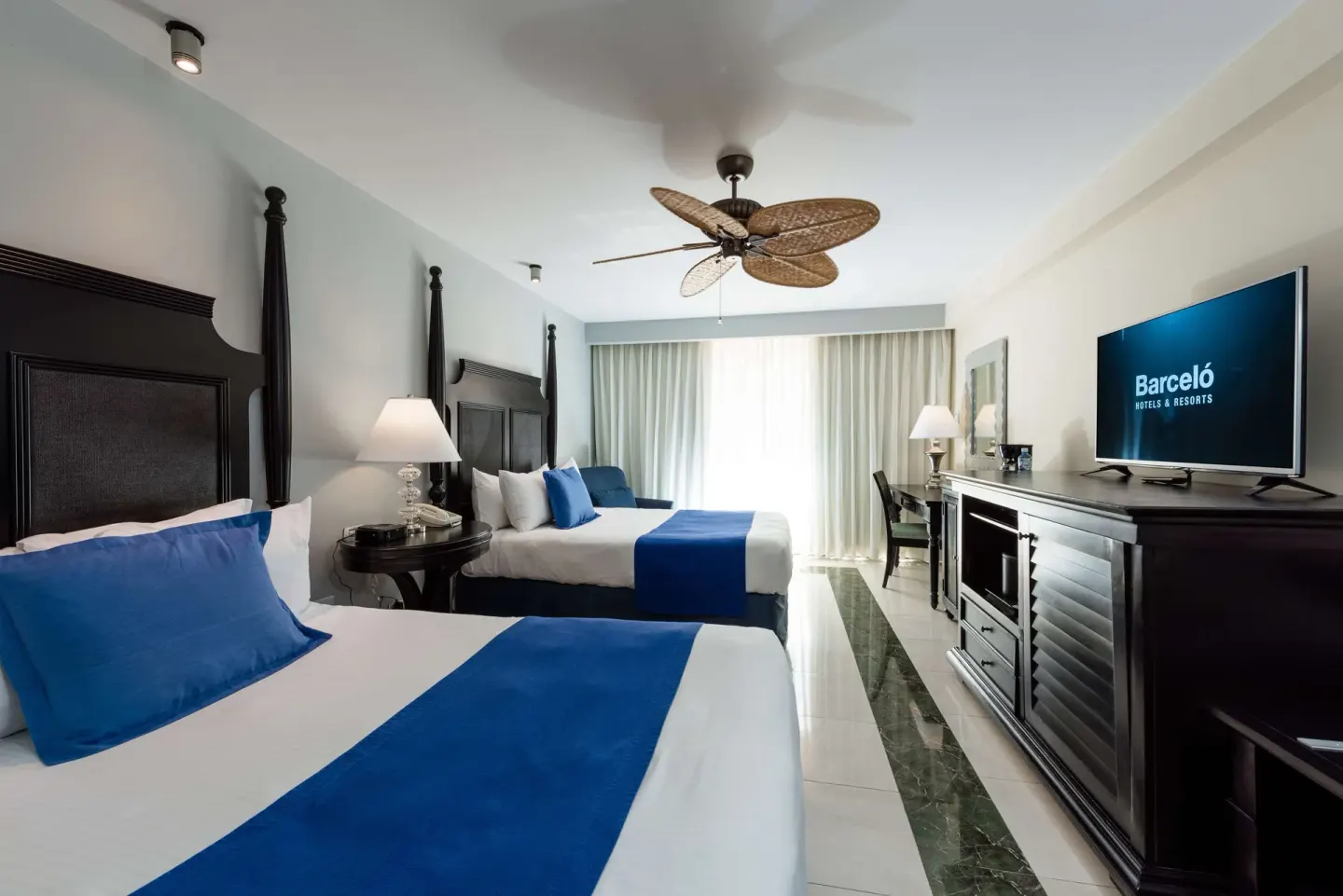 Double-bed guest room with TV, dresser, desk, and balcony curtains at Barceló Aruba.