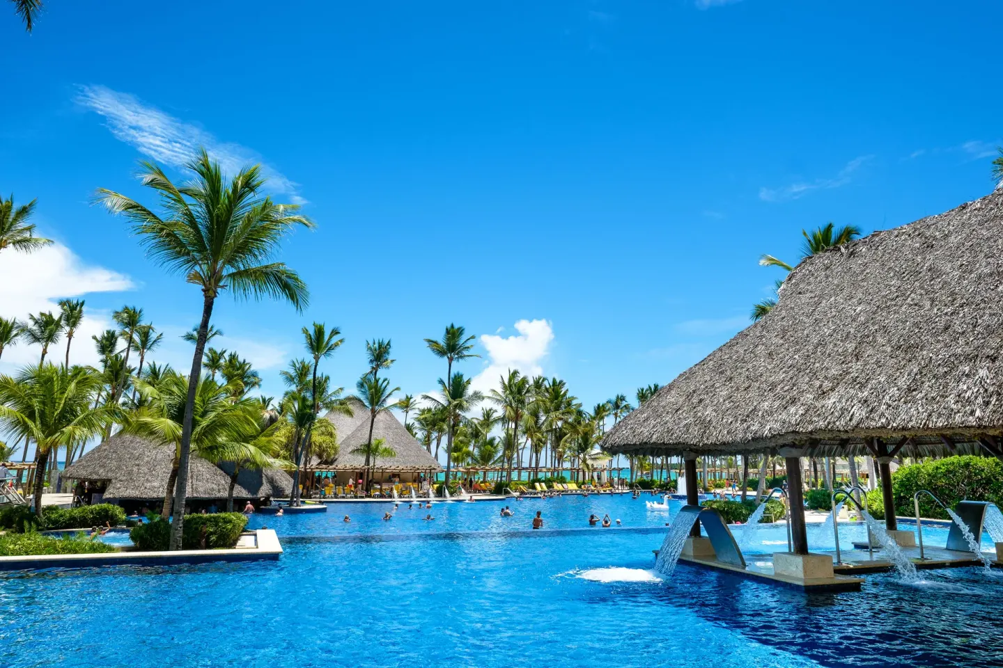 Large resort pool with thatched shade structures and palm trees at Barceló Bávaro Palace.