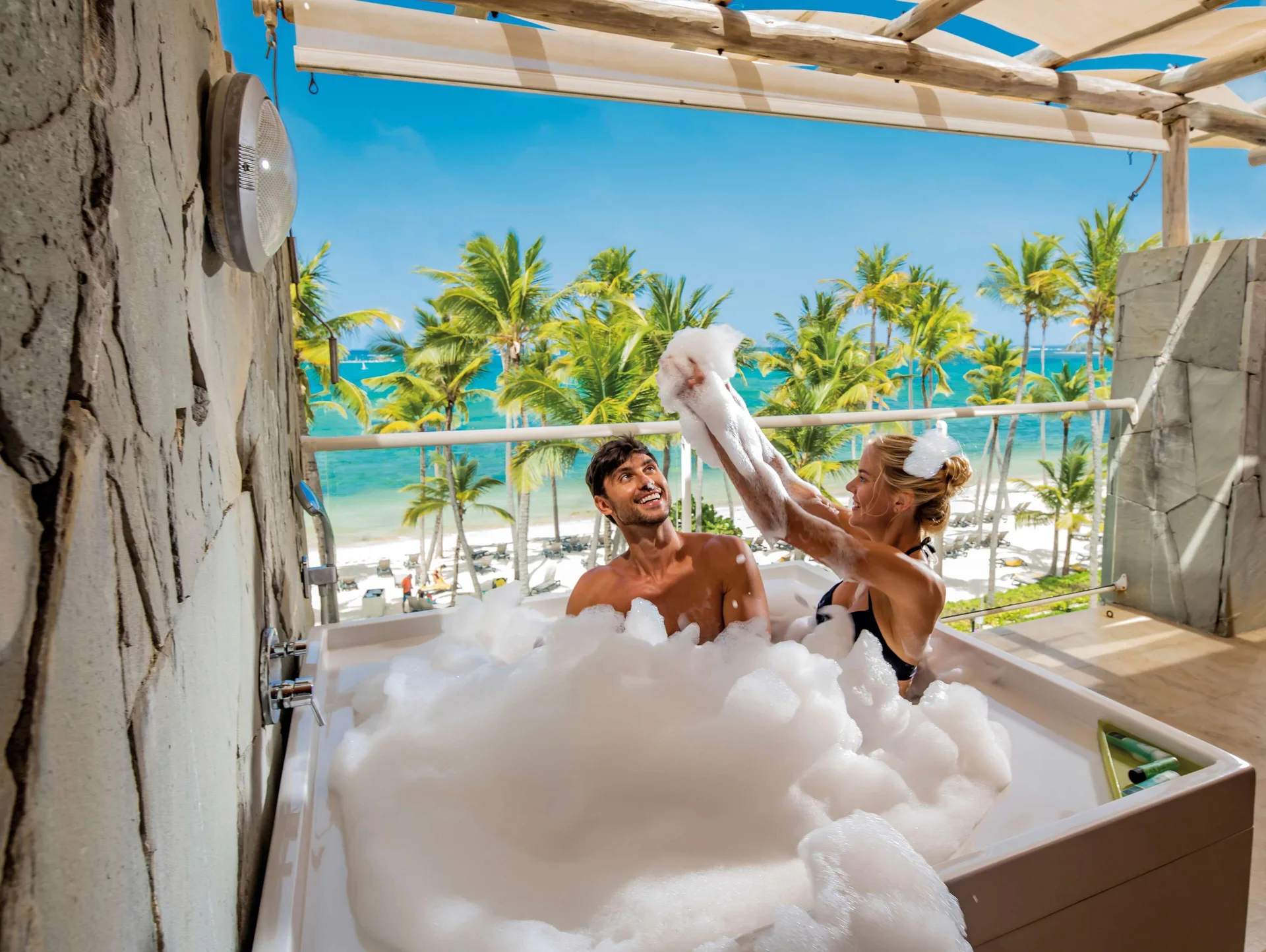 Couple in a foamy balcony tub with ocean and palms at Barceló Bávaro Palace.