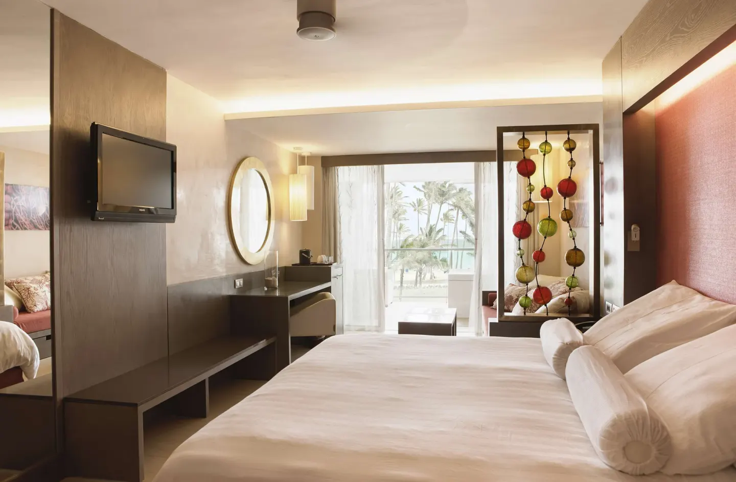 Junior Suite king bed with desk, wall TV, decorative screen, and ocean-facing balcony at Barceló Bávaro Palace.